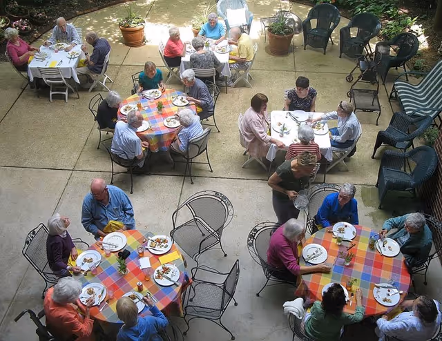 A group of elderly people seated at round tables with colorful checkered tablecloths, enjoying a meal together outdoors on a patio surrounded by potted plants and additional empty chairs.