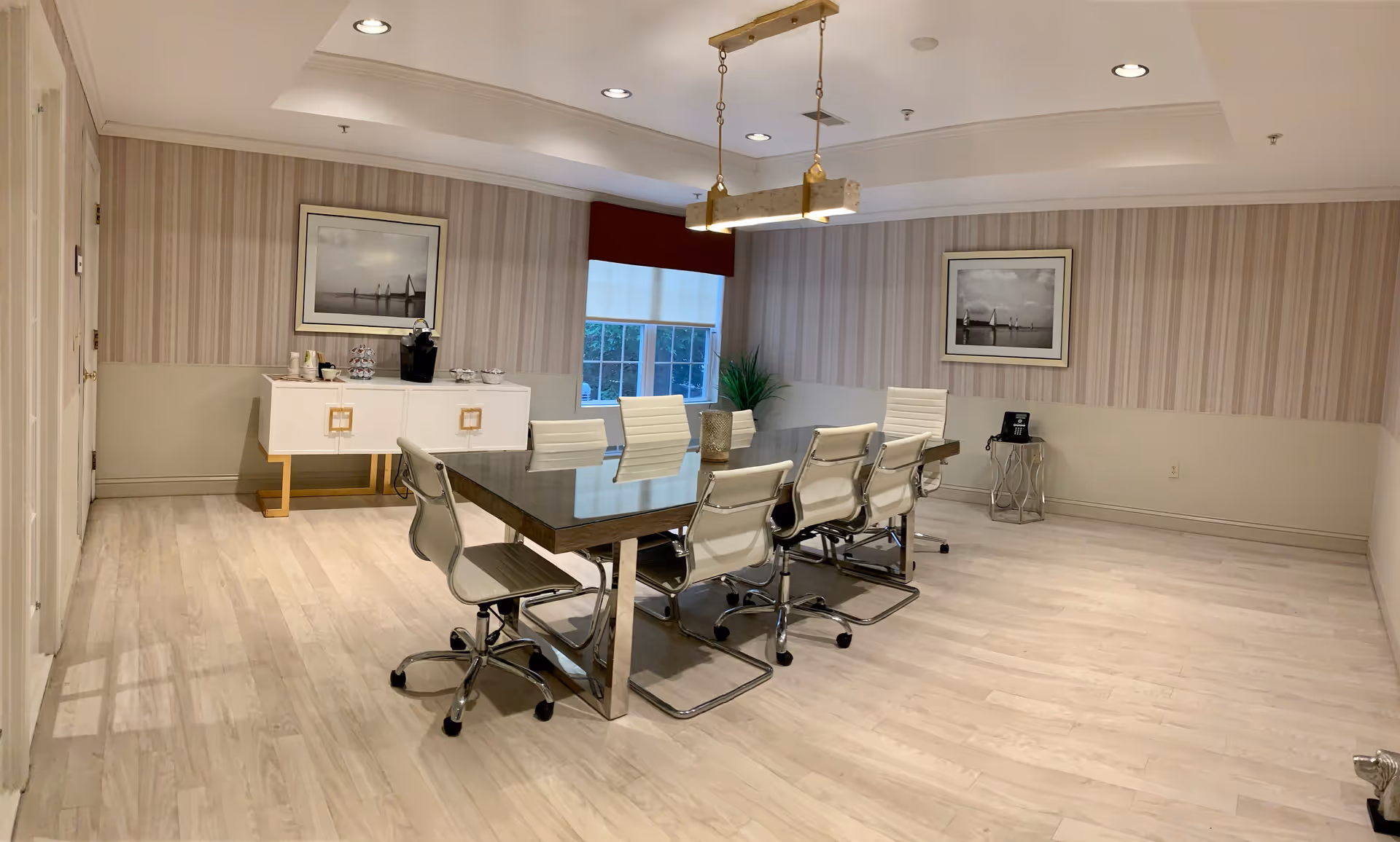 A modern conference or meeting room with a long rectangular table surrounded by eight white office chairs. The room has light wood flooring, striped wallpaper, and two framed black and white pictures on the walls. A white sideboard with coffee supplies is against one wall, and a small side table with a phone is in the corner. A window with a red valance and white blinds lets in natural light. Two rectangular pendant lights hang from the ceiling above the table.