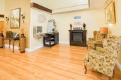 Small common room with hardwood floors, two floral armchairs, a fireplace, and a coffee/coffee station against a cream wall.