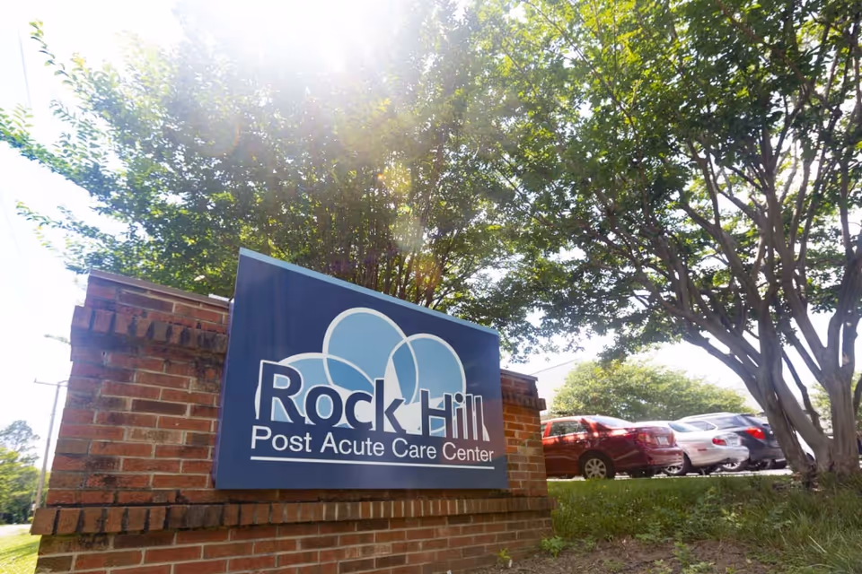 Outdoor view of a brick sign for Rock Hill Post Acute Care Center with trees and parked cars in the background under a bright sunny sky.