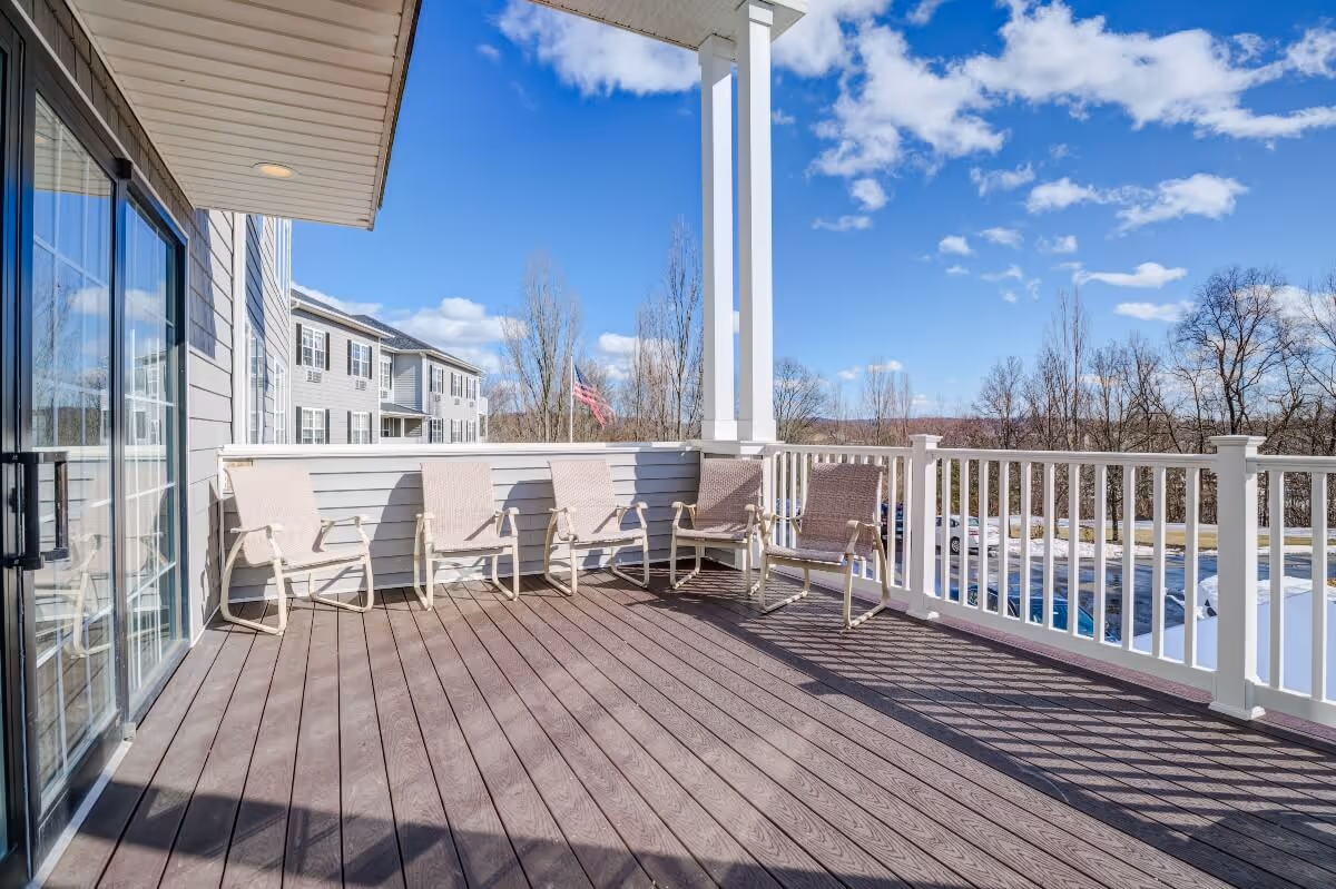 Outdoor balcony area with five beige chairs arranged along the railing. The balcony has wooden flooring and white railings, with a view of a parking lot, trees, and a partly cloudy blue sky.