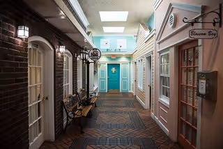 Indoor hallway designed to resemble an outdoor street with small storefront-style doors and windows on either side. The floor is made of brick tiles, and there are benches along the left side. The ceiling has skylights allowing natural light to enter. Signs above the doors read 'Blossom Bay' and other names, creating a quaint village atmosphere.