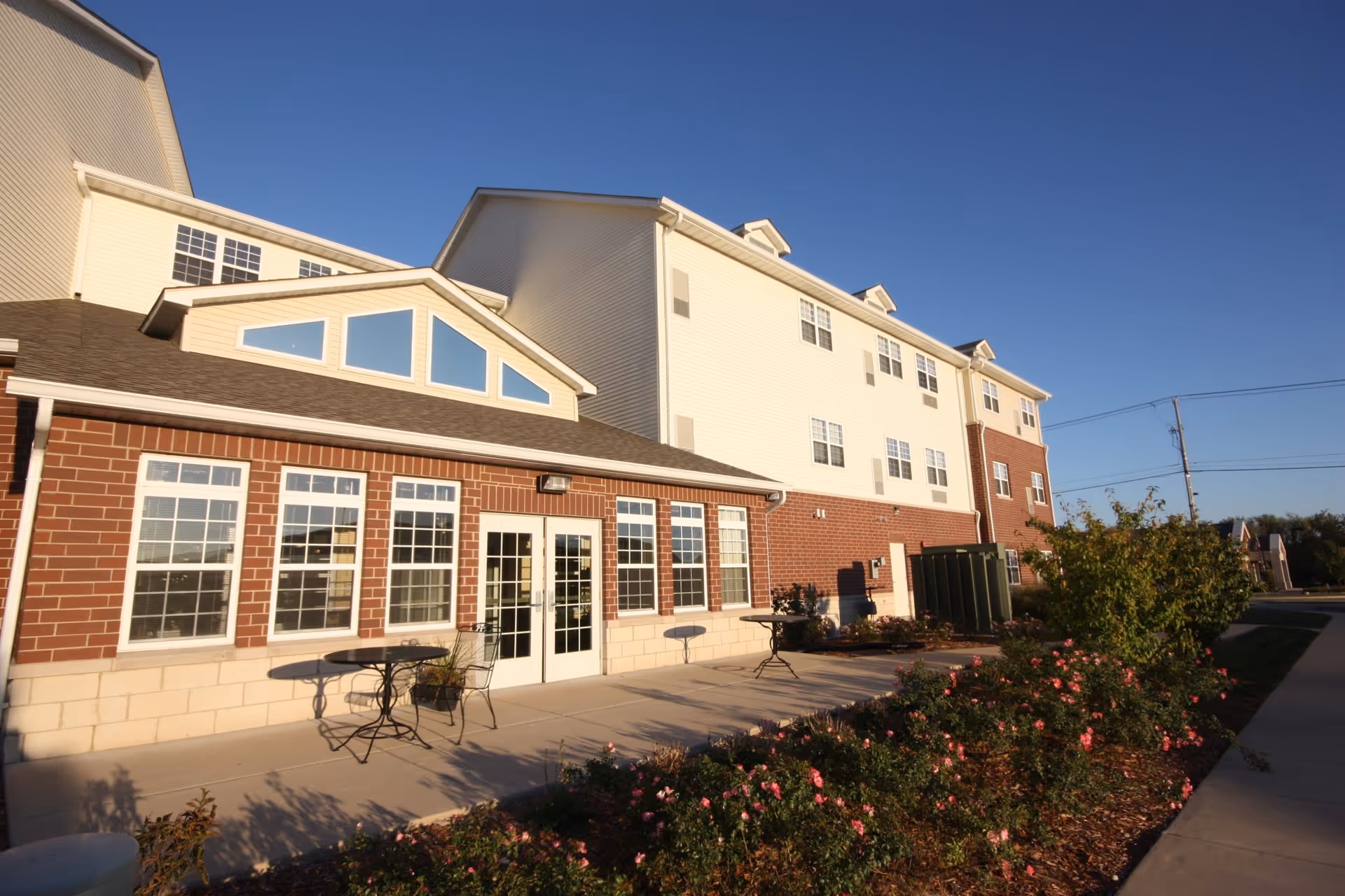 Exterior view of a multi-story senior living facility building with a brick and light-colored siding facade. The building has multiple windows and a patio area with metal tables and chairs. There are flower beds with blooming pink flowers along the sidewalk in front of the building under a clear blue sky.