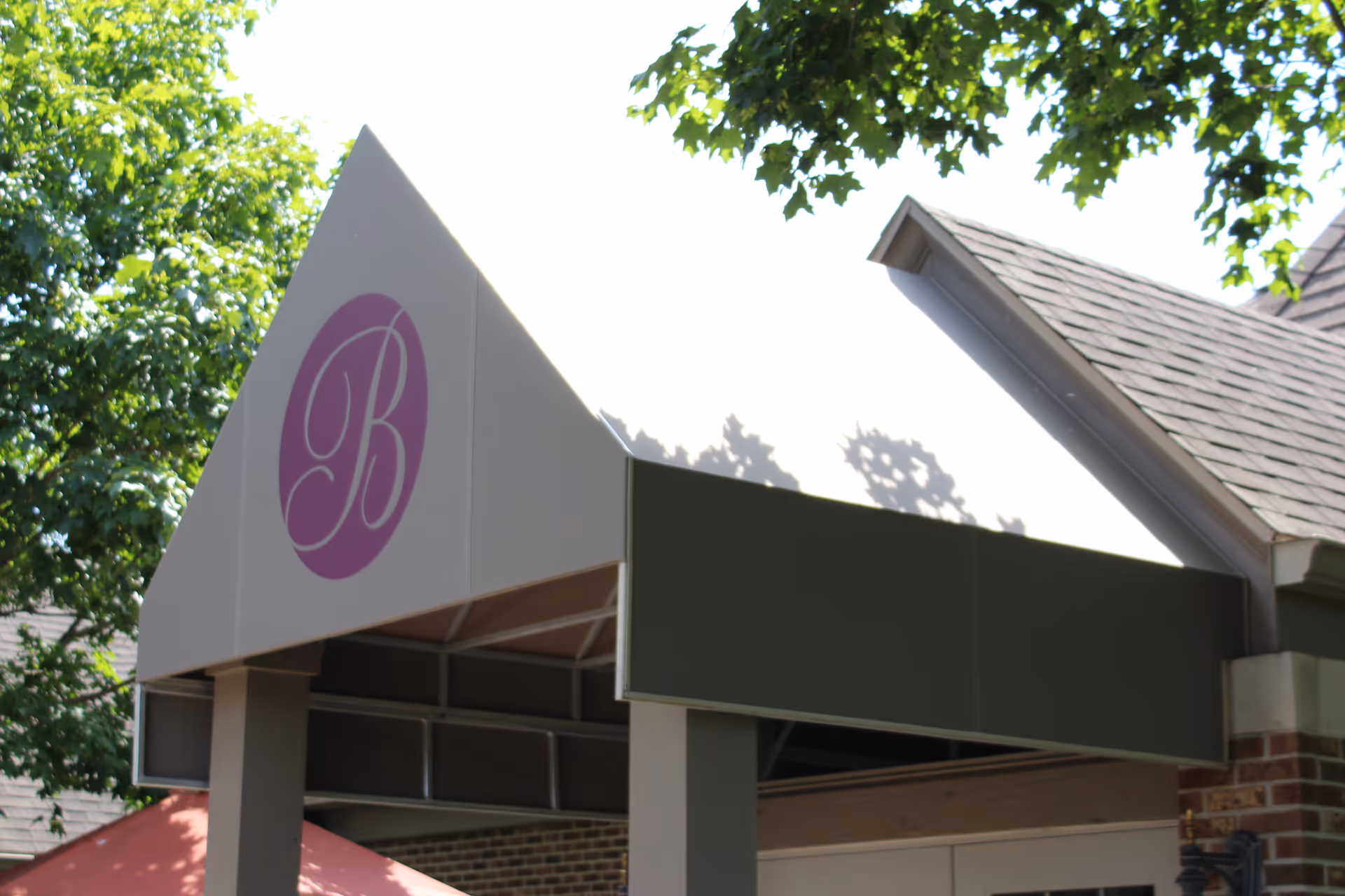 Entrance canopy of a building with a triangular roof structure featuring a purple circle with a white letter 'B' on it, surrounded by green trees and part of a brick wall and shingled roof visible.