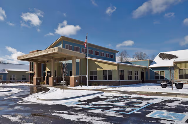 Exterior view of The Springs of Mooresville facility on a snowy day, showing a modern building with a covered entrance, an American flag on a pole, and a partially snow-covered parking lot with handicap parking spaces.
