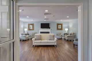 View through a doorway into a living room area with a central beige sofa, multiple armchairs arranged around the room, a fireplace with a mounted flat screen TV above it, two framed pictures on the wall, and lamps on side tables providing soft lighting. The floor is wood and the walls are painted light blue.