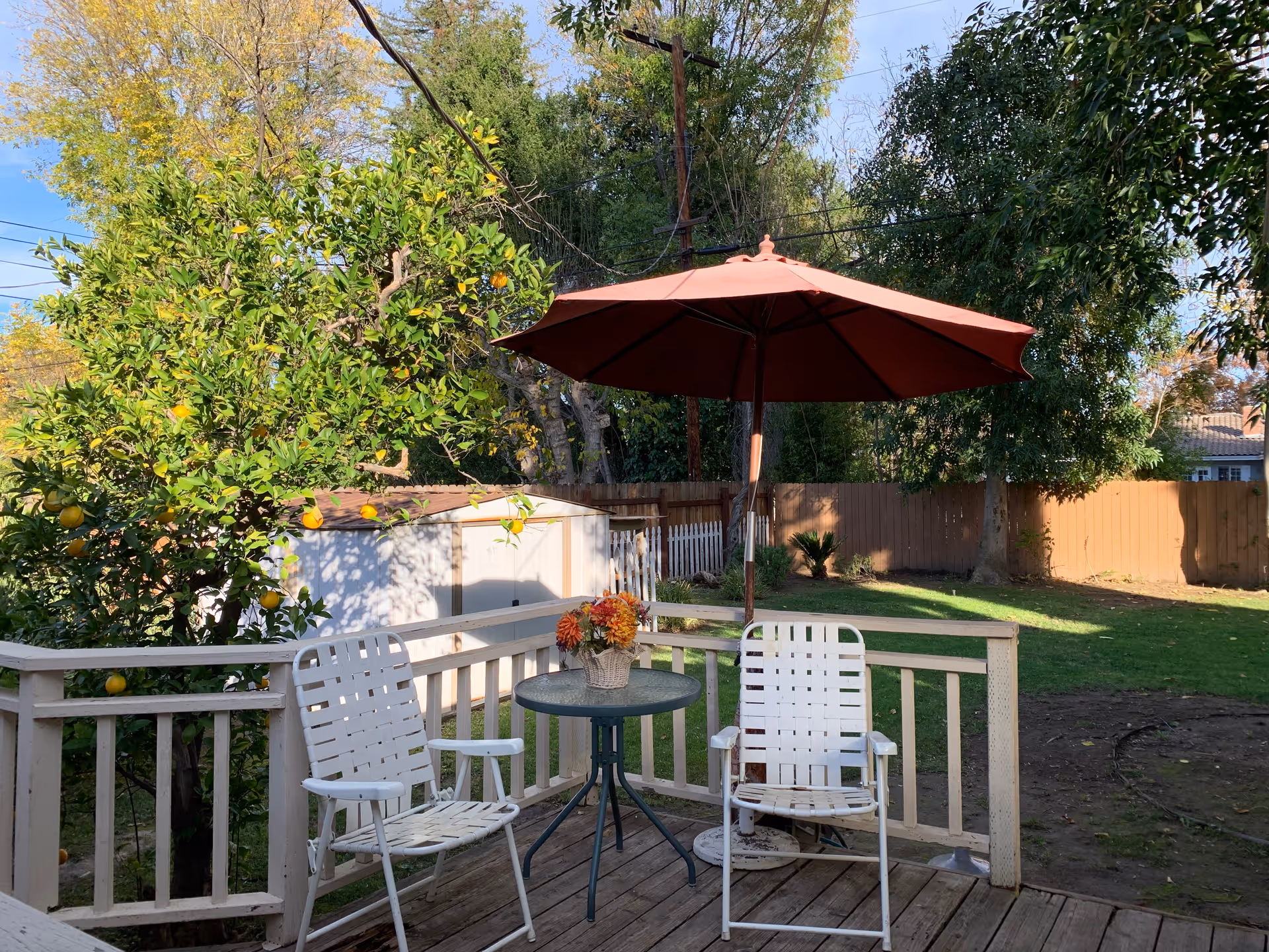 A backyard patio area with two white metal chairs and a small round glass table with a flower arrangement on it. A large red umbrella provides shade. The patio is surrounded by a white wooden railing. In the background, there is a green lawn, trees, a wooden fence, and a small white shed. The sky is clear and blue.