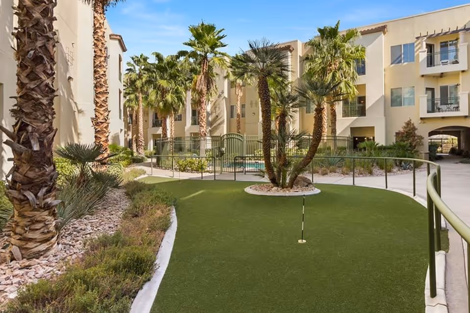 Outdoor courtyard area at Merrill Gardens at Green Valley Ranch featuring a putting green surrounded by palm trees and desert landscaping, with multi-story beige residential buildings in the background under a clear blue sky.