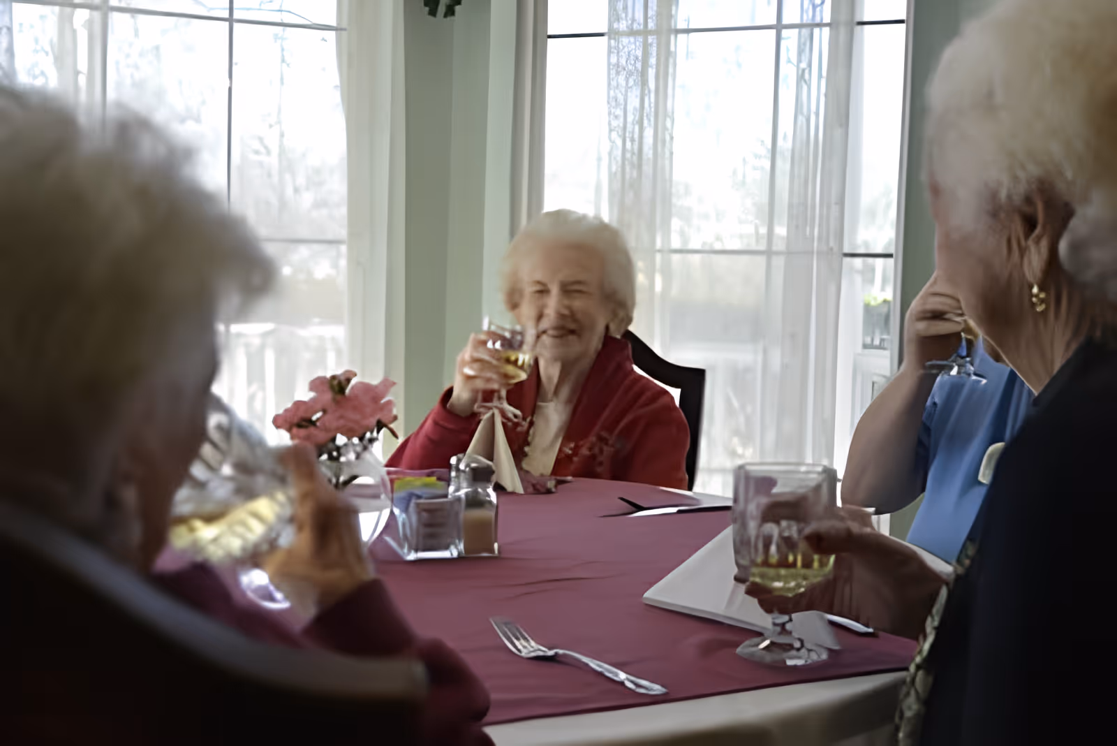 A group of elderly women sitting around a dining table with a maroon tablecloth, holding glasses and enjoying a drink together in a bright room with large windows and sheer curtains.