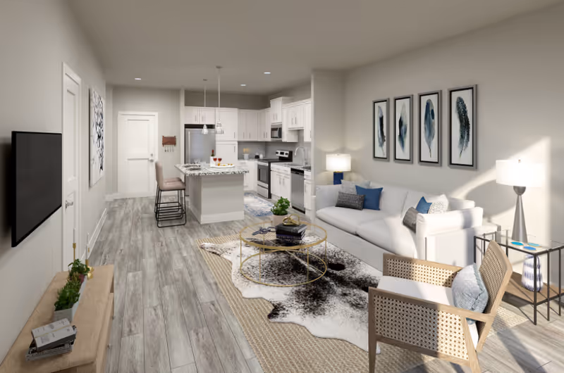 A modern open-concept living room and kitchen area with light wood flooring. The living room features a white sofa with blue and gray cushions, a wooden chair with a cushion, a round glass coffee table on a patterned rug, and framed artwork of feathers on the wall. The kitchen has white cabinets, stainless steel appliances, a small island with two bar stools, and pendant lights hanging above the island.