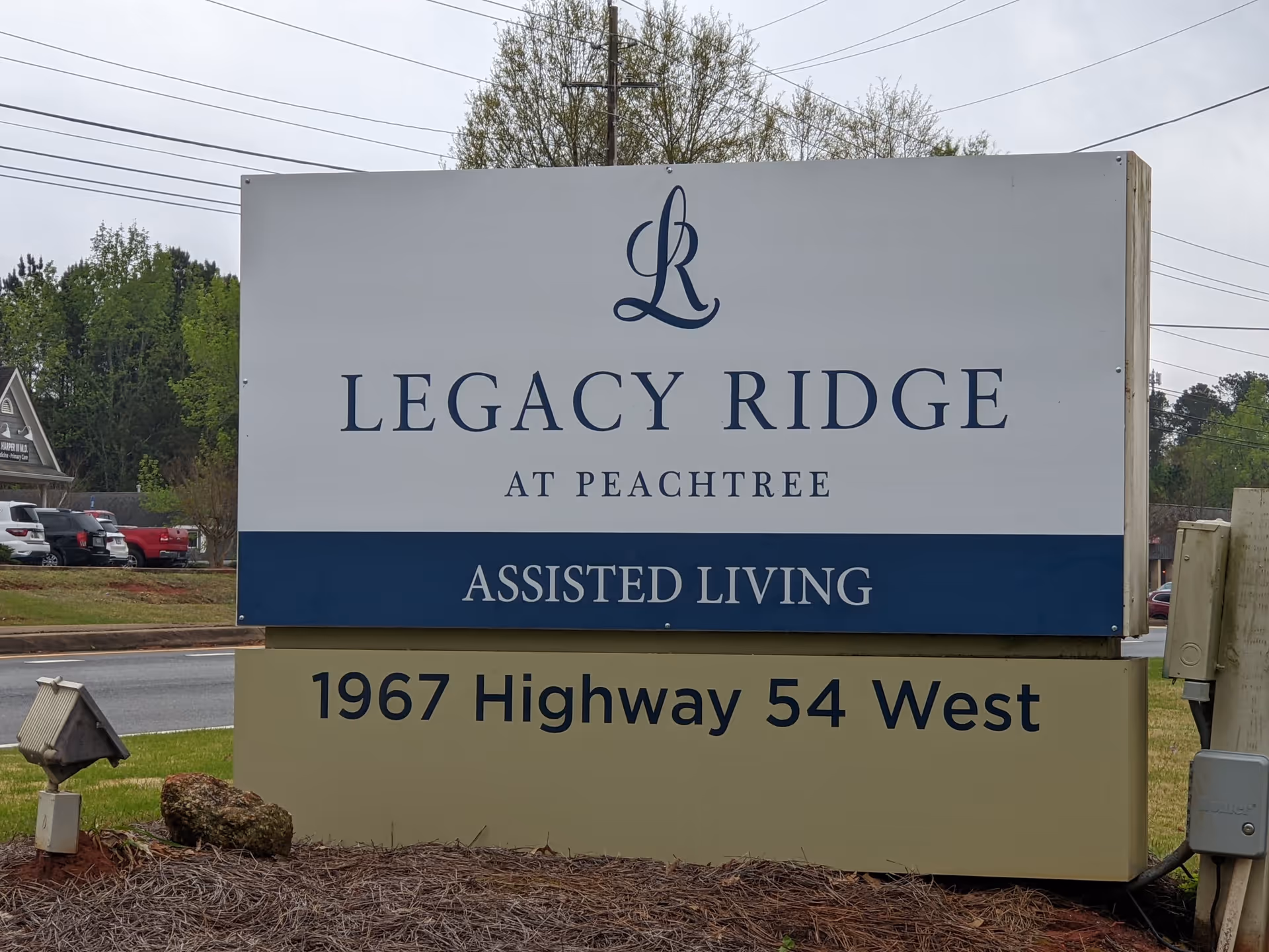 Outdoor sign for Legacy Ridge at Peachtree, an assisted living facility, located at 1967 Highway 54 West. The sign is white and blue with black text, set on a beige base with grass and a small rock in front.