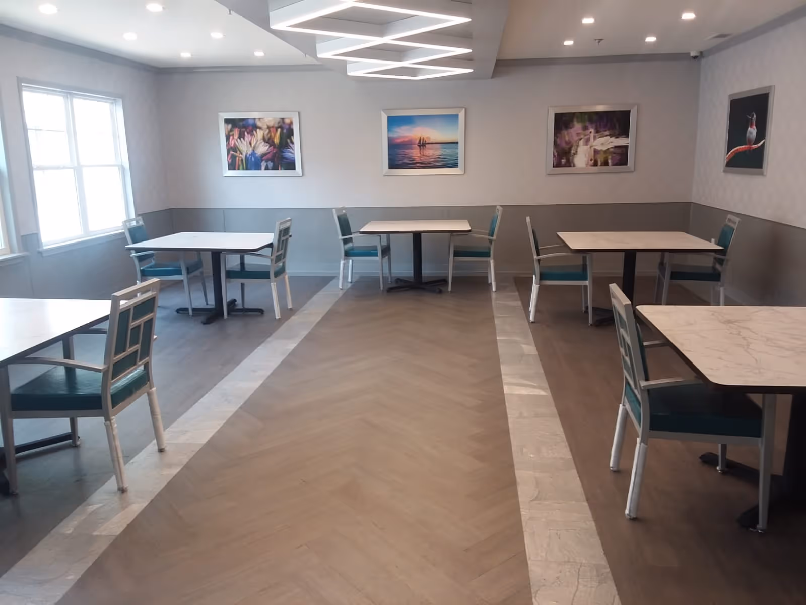 Communal dining room with multiple small square tables and teal-upholstered chairs, artwork on the walls and modern overhead lighting.