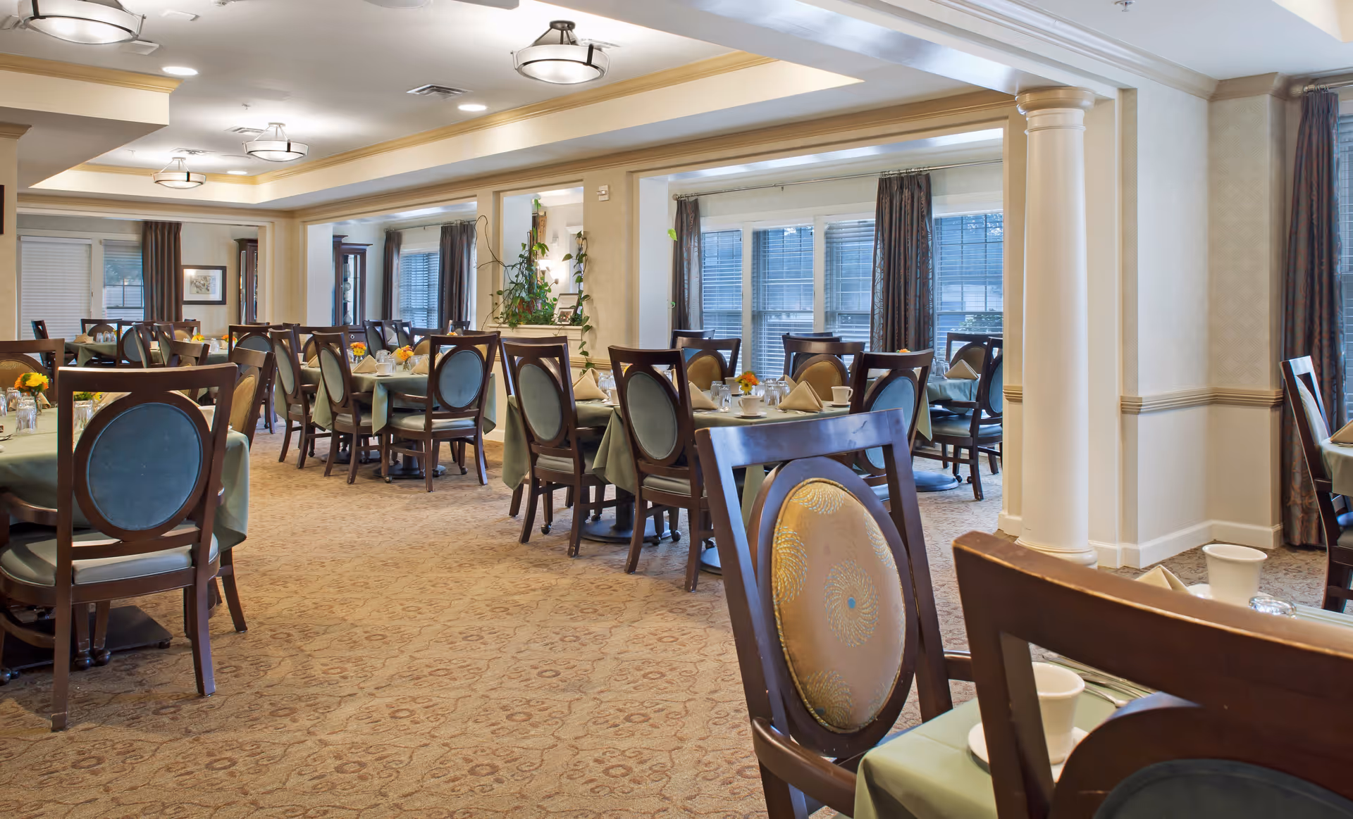 Spacious dining room with multiple set tables and upholstered chairs, columns, and large windows.