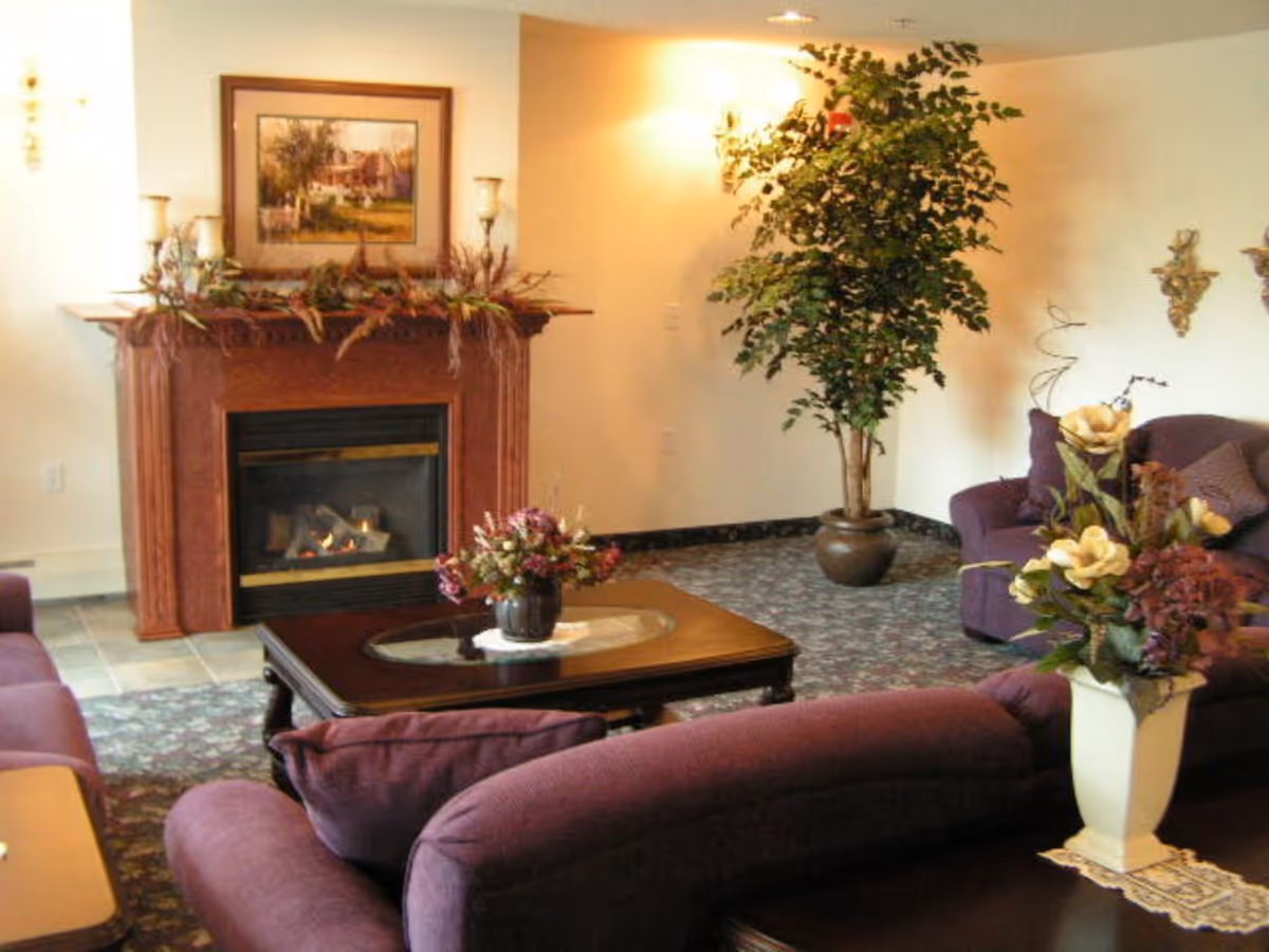 A cozy living room with purple upholstered sofas arranged around a wooden coffee table with a floral centerpiece. There is a fireplace with a wooden mantle decorated with candles and greenery, and a framed painting above it. A large potted plant is placed in the corner, and the room has soft lighting and floral carpet.