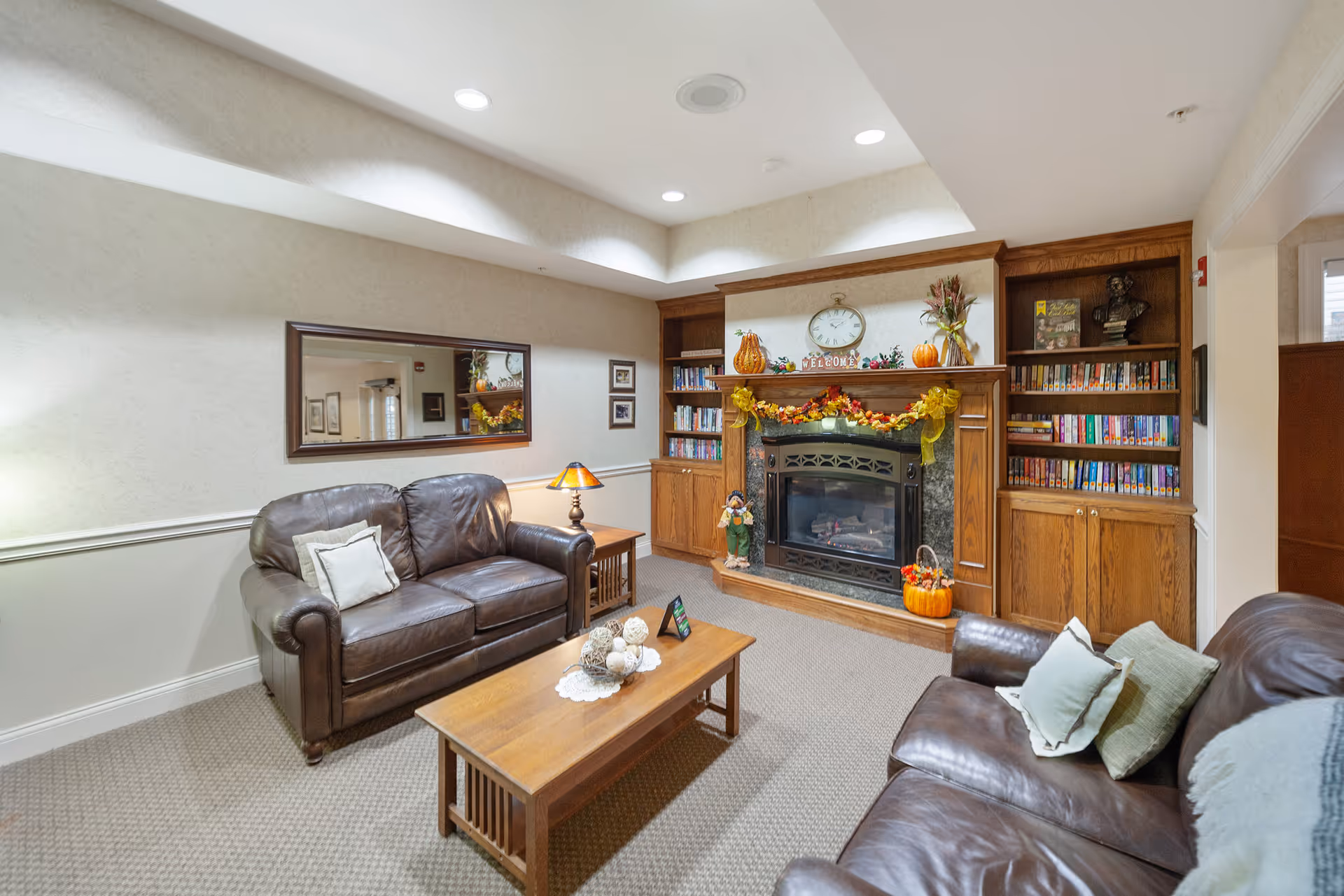 A cozy living room with two brown leather sofas facing a wooden coffee table. The room features a fireplace decorated with autumn-themed garlands, pumpkins, and a clock above the mantel. Built-in wooden bookshelves flank the fireplace, filled with books. A large mirror hangs on the wall above one sofa, and a small table with a lamp is positioned between the sofas. The room has soft lighting and a carpeted floor.
