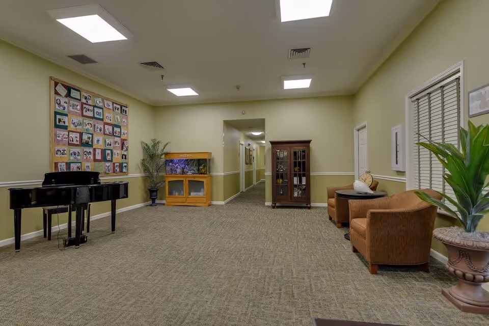 Interior view of a senior living facility common area with a black grand piano on the left, a colorful quilt with photos on the wall, a fish tank on a wooden stand, a wooden cabinet, two brown armchairs with a small round table between them, and potted plants. The walls are light green with white trim, and the ceiling has recessed lighting.