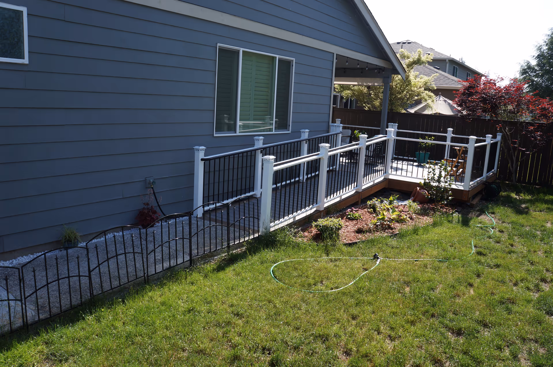 Backyard view of a gray house with a raised deck and white railing, small garden beds and a grassy lawn.