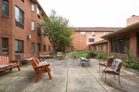 Outdoor courtyard area at Independence Village of East Lansing featuring concrete walkways, several wooden and metal chairs with cushions, benches, and surrounding brick buildings with windows. There are some trees and plants along the edges of the courtyard.