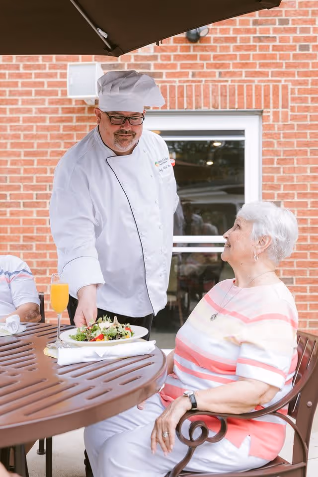 A chef in a white uniform and hat serves a plate of salad to an elderly woman seated at an outdoor table. The woman is smiling and looking up at the chef. The setting is outside with a brick wall and window in the background.
