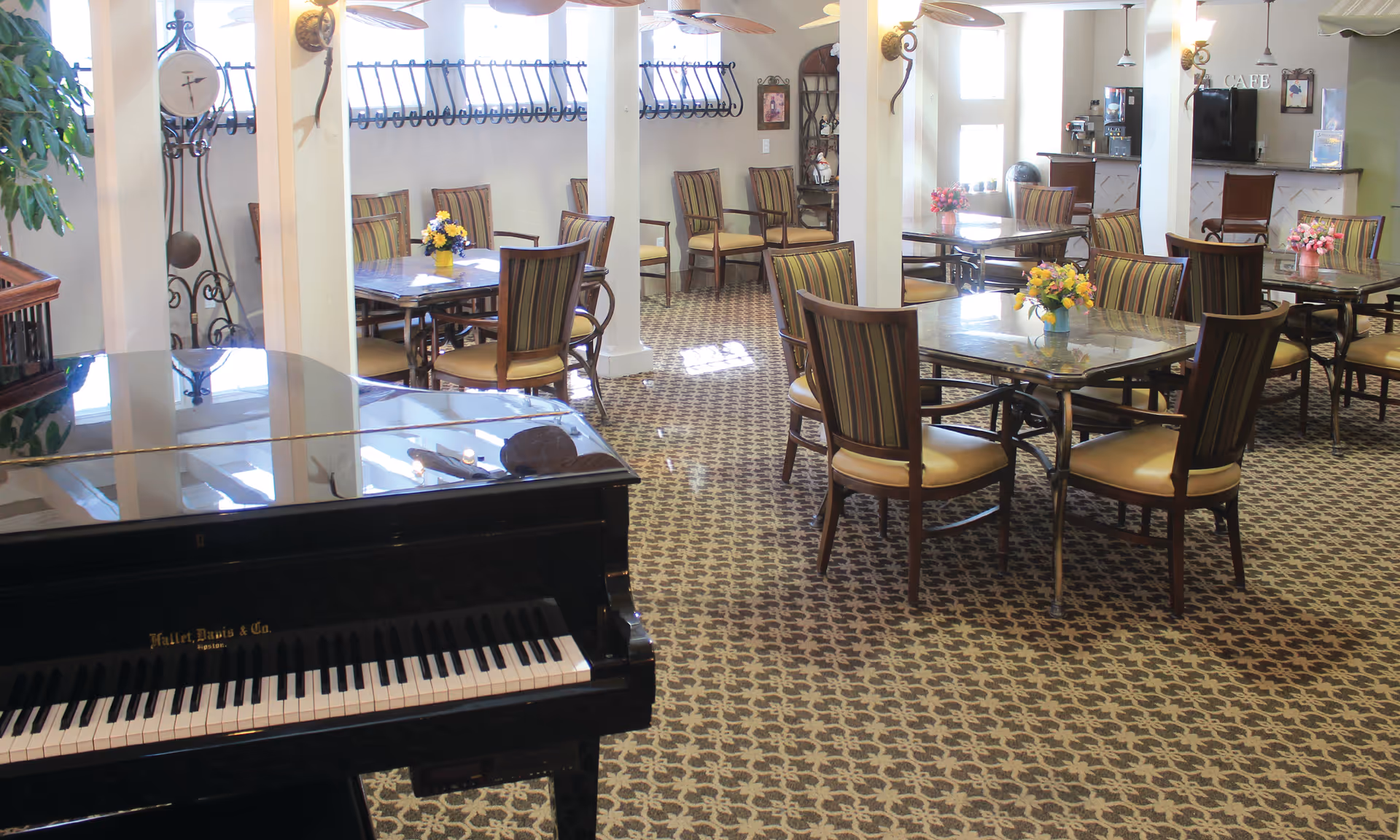 Bright senior living dining area with round tables and chairs, floral centerpieces, and a piano in the foreground.