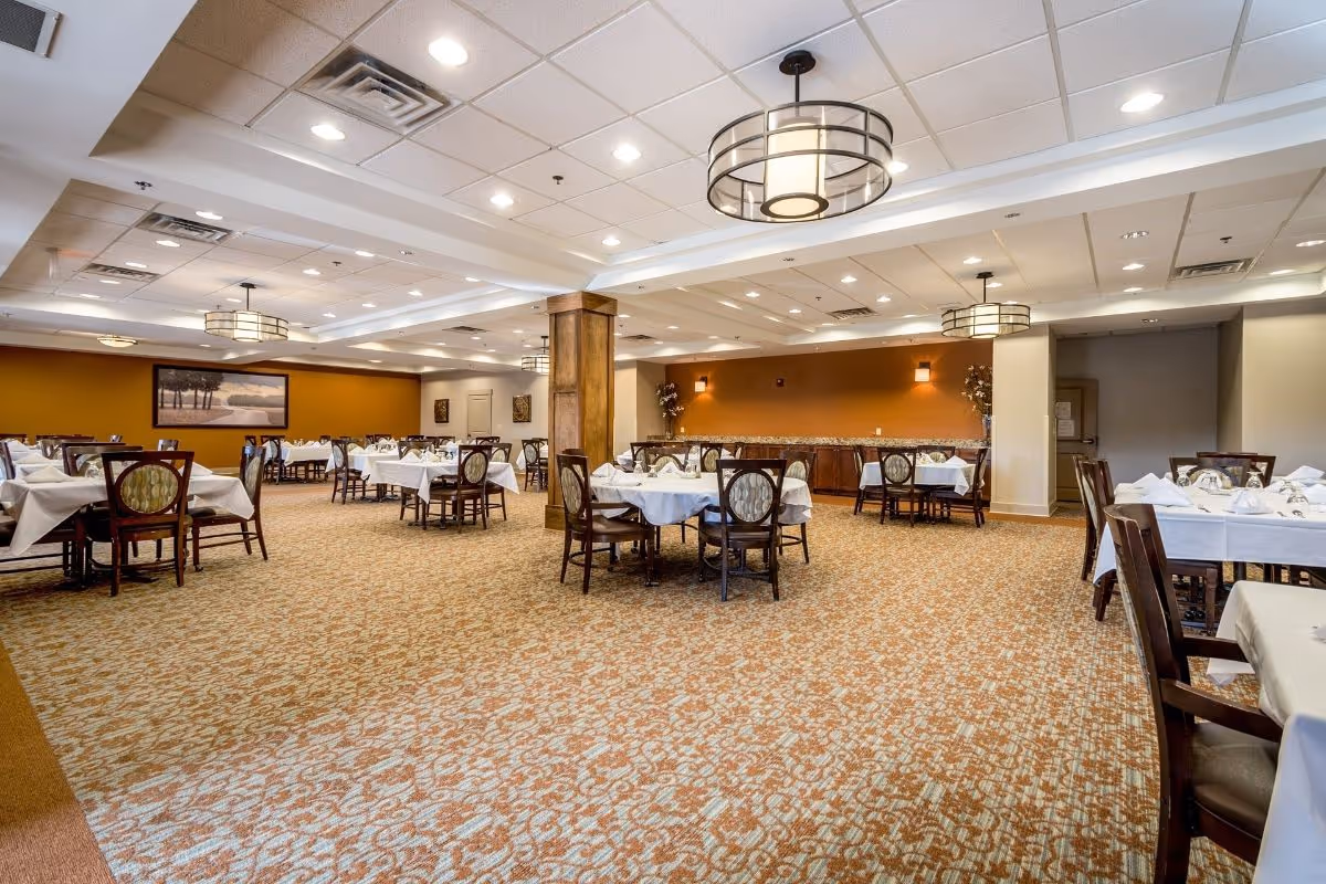 Spacious dining room with multiple tables covered in white tablecloths and set with glassware and napkins. The room features patterned carpet, warm orange accent walls, wooden chairs with cushioned seats, ceiling lights with circular metal frames, and framed artwork on the walls.