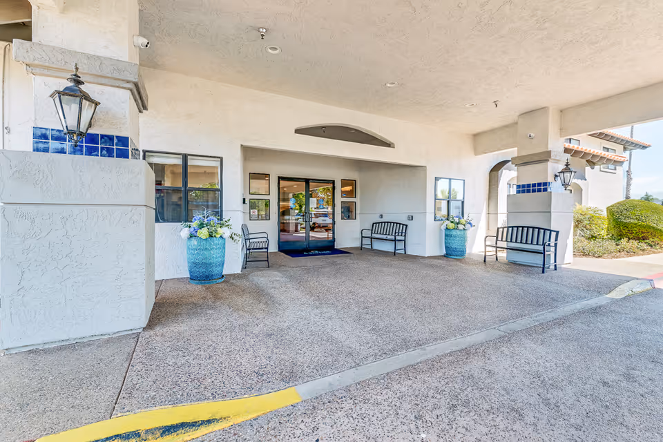 Covered entrance area of Solstice Senior Living at El Cajon with a textured concrete floor, white stucco walls, two large blue flower pots with floral arrangements, black metal benches, and glass double doors leading inside.