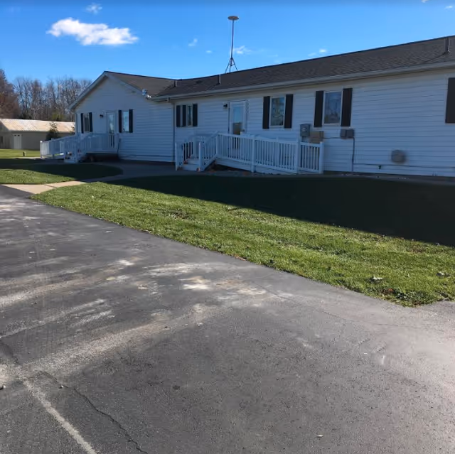 Exterior view of a single-story white building with a sloped roof, multiple windows with dark shutters, and two white ramps leading to separate entrances. The building is surrounded by a green lawn and a paved driveway under a blue sky with a few clouds.