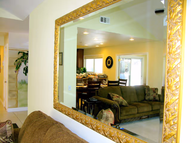 A large ornate gold-framed mirror reflecting a living room and dining area. The living room has a brown sofa with patterned cushions, and the dining area has a wooden table with chairs. There is a sliding glass door in the background letting in natural light.