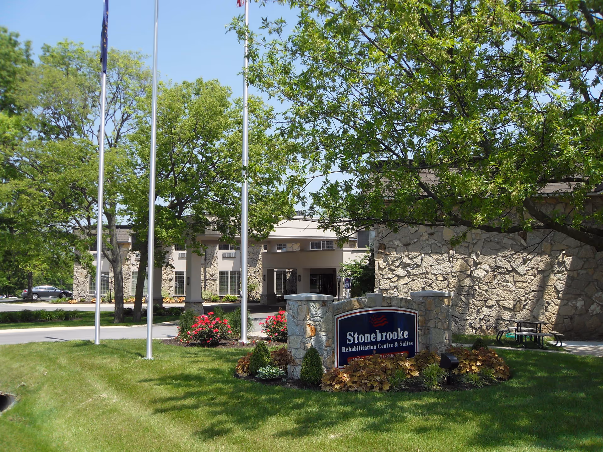Front exterior of Stonebrooke Rehabilitation Center showing the entrance, a stone identification sign, flagpoles, trees, and a landscaped lawn.