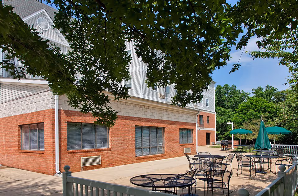 Outdoor patio area at Woodside Village featuring several round metal tables with chairs and green umbrellas. The patio is adjacent to a brick and white siding building, surrounded by trees and greenery under a clear blue sky.
