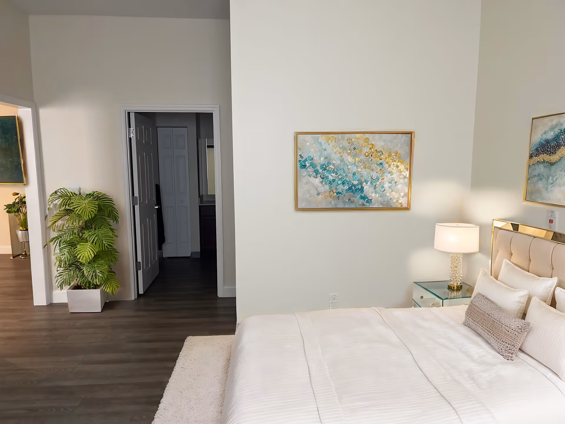 Bright, modern bedroom with a neatly made bed, nightstand and lamp, abstract wall art, and an open doorway to a hallway.