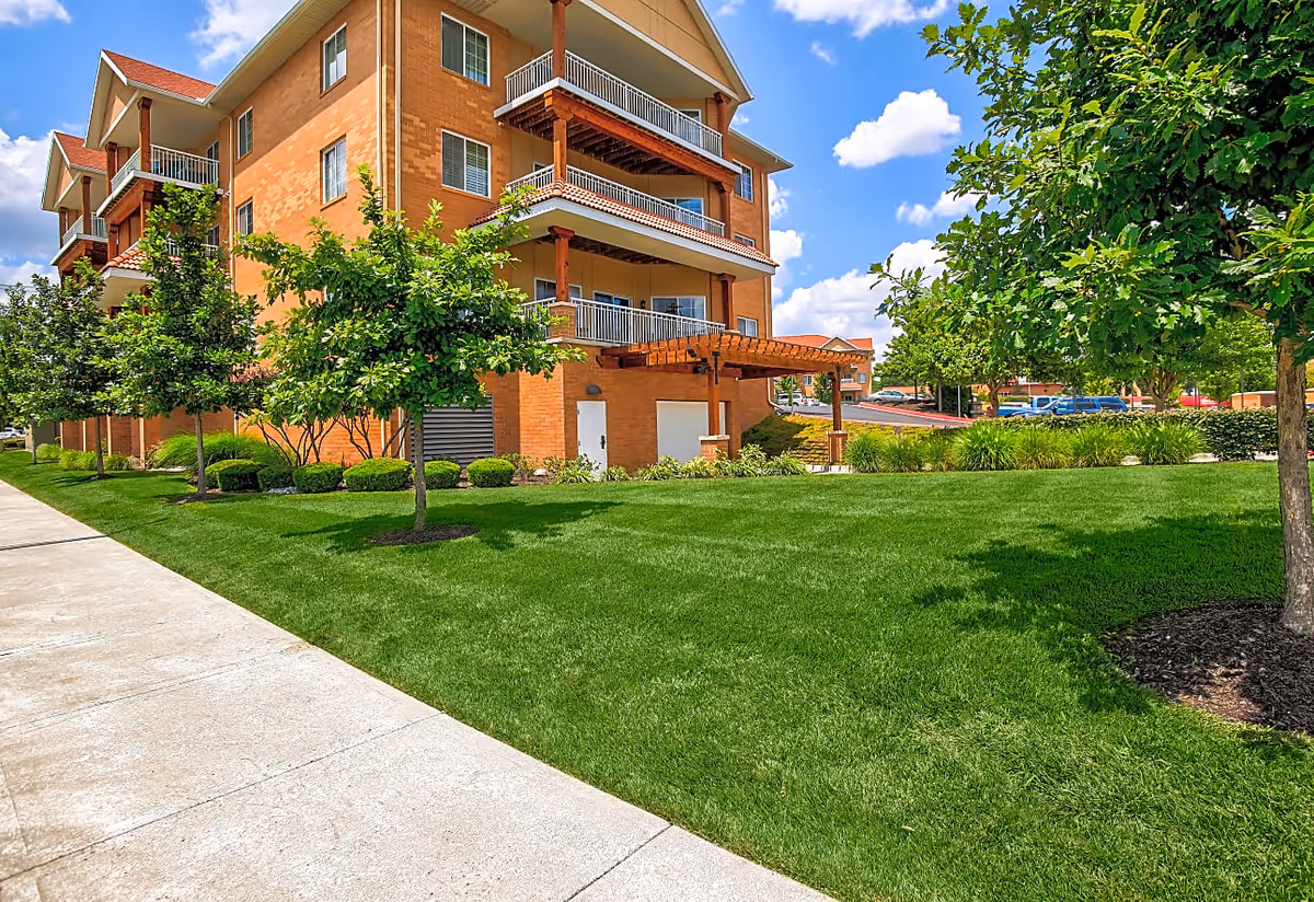 Exterior view of a multi-story brick senior living building with balconies, a green lawn, and trees.