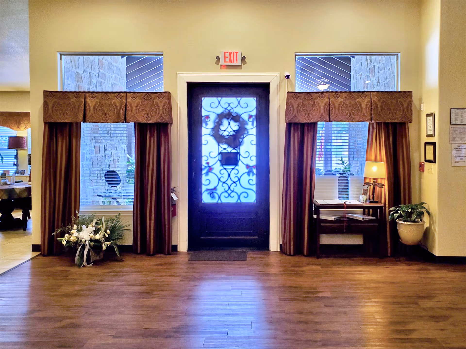 Interior view of an assisted living facility entrance area with a decorative glass door in the center, flanked by two windows with brown curtains and valances. There is a floral arrangement on the left side and a small table with a lamp, books, and a plant on the right side. The floor is wooden, and there is an exit sign above the door.