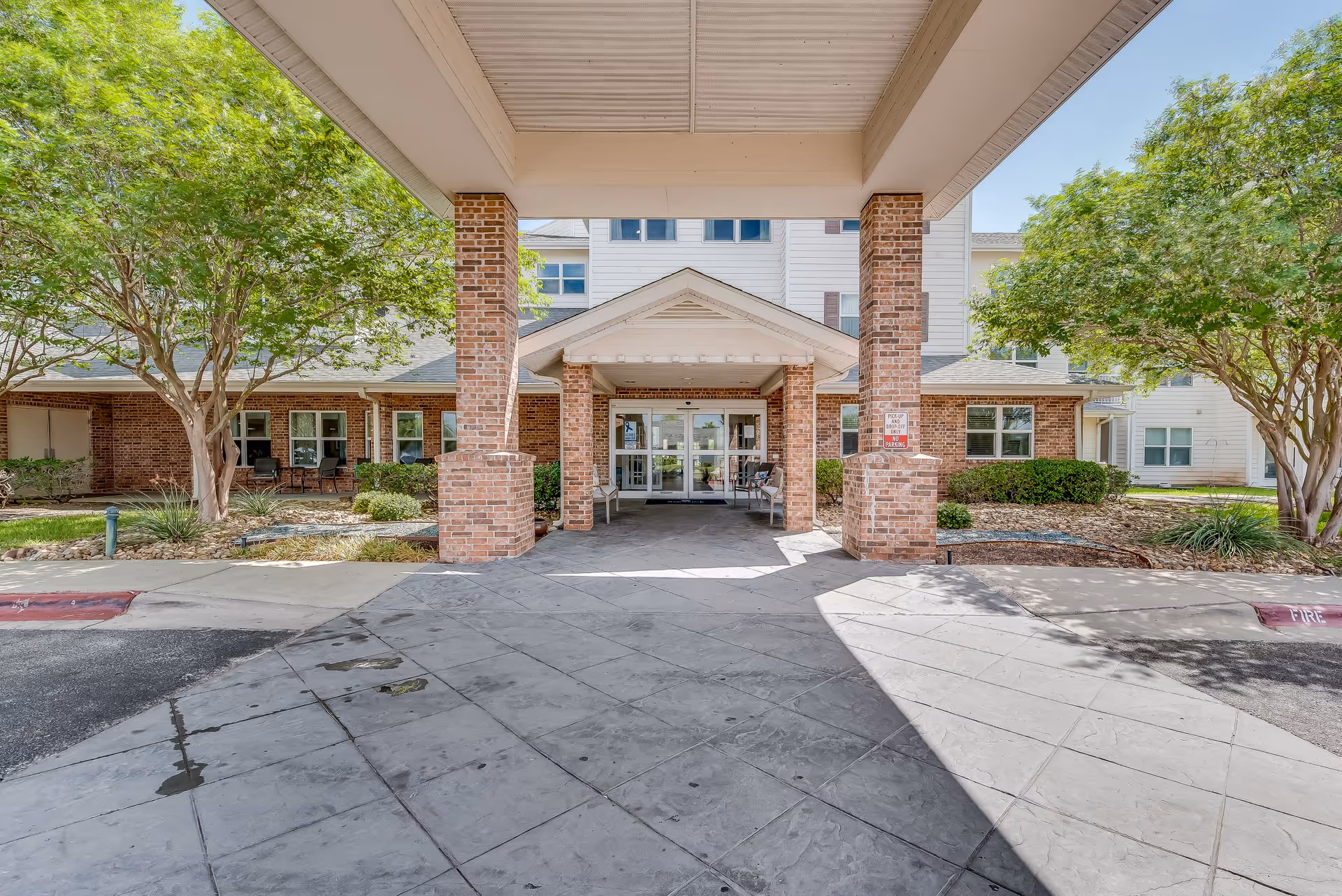 Entrance to a senior living facility with a covered drop-off area supported by brick columns. The building features a combination of brick and white siding, with large windows and a glass door entrance. There are trees and landscaping on either side of the entrance.