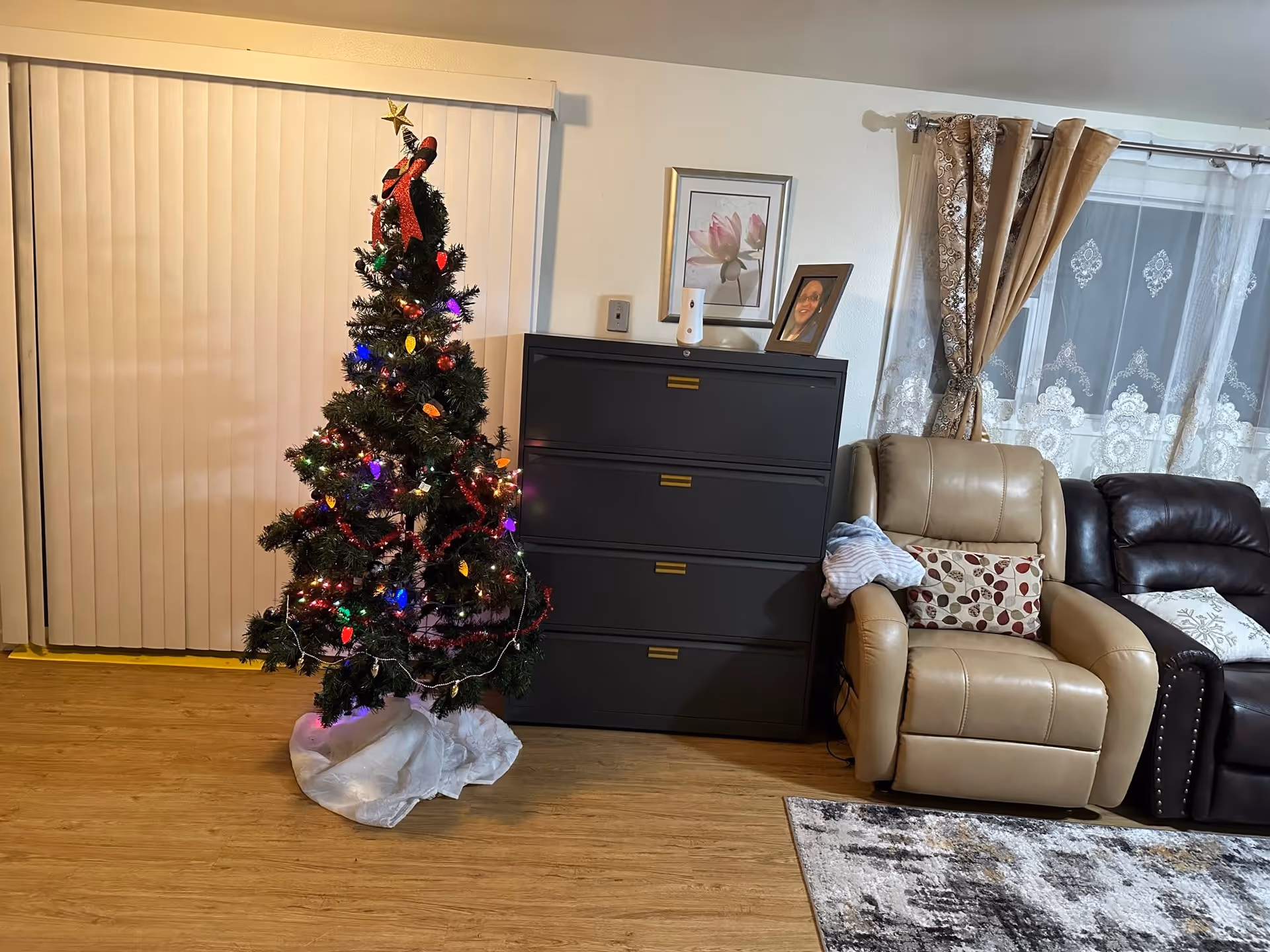 A decorated Christmas tree with colorful lights and ornaments stands next to a black filing cabinet with a framed photo and a small device on top. To the right, there is a beige recliner chair with a patterned pillow and a folded blanket on the armrest, next to a dark brown leather sofa. The room has wooden flooring, vertical blinds on the left, and a window with sheer and patterned curtains on the right.
