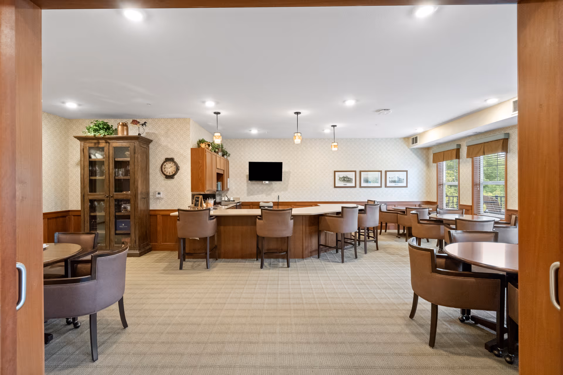 A spacious dining area with multiple round tables and brown cushioned chairs. The room features a central counter with high chairs, pendant lights hanging from the ceiling, a wall-mounted TV, and framed pictures on the wall. Large windows on the right side allow natural light to enter, and a wooden cabinet with glass doors is visible on the left side.