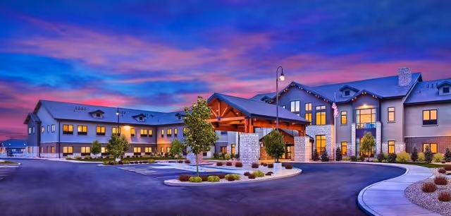 Exterior view of Copper Creek Assisted Living & Memory Care building at dusk with a colorful purple and pink sky. The building is two stories with warm lights glowing from the windows, a covered entrance with wooden beams, landscaped greenery, and a curved driveway in front.