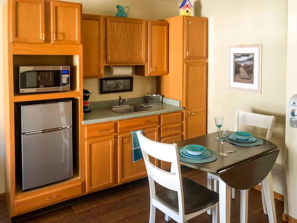 A small kitchen area with wooden cabinets, a stainless steel microwave and mini refrigerator, a sink with a countertop, and a small dining table set for two with white chairs and teal dishware. A framed picture hangs on the wall and a hand sanitizer dispenser is mounted nearby.