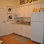 A small kitchen area with white cabinets, a white refrigerator, a microwave above the countertop, and a sink. There are two framed pictures on the wall to the left, a small rug on the wooden floor in front of the sink, and various kitchen items on the countertop.