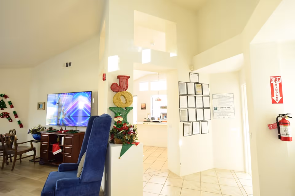 Bright common room interior with a television, blue armchair, dining chairs, holiday decorations, and framed notices on the wall.