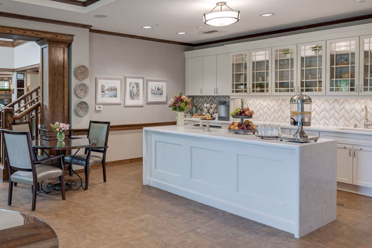 A bright and clean kitchen area in a senior living facility with a large white island countertop. The countertop holds a tiered fruit tray, a water dispenser, glasses, and a vase with flowers. Behind the island are white cabinets with glass doors displaying dishes and decor. To the left, there is a small round table with four chairs and a vase of flowers. The walls are decorated with framed artwork and decorative plates, and there is wood trim around the room.