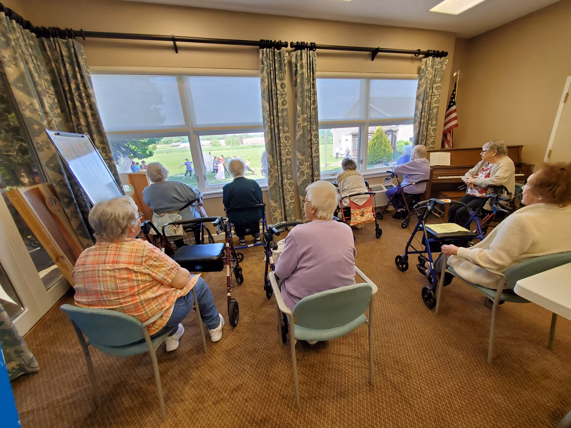 A group of elderly people sitting in chairs and using walkers in a room with large windows looking out onto a green outdoor area. The room has patterned curtains, a piano with an American flag beside it, and a whiteboard on an easel. The elderly individuals appear to be watching an event or activity outside.