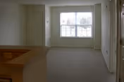 Empty interior room with beige carpet, a large window letting in natural light, a closed door on the left wall, and a partial view of a wooden railing or countertop in the foreground.