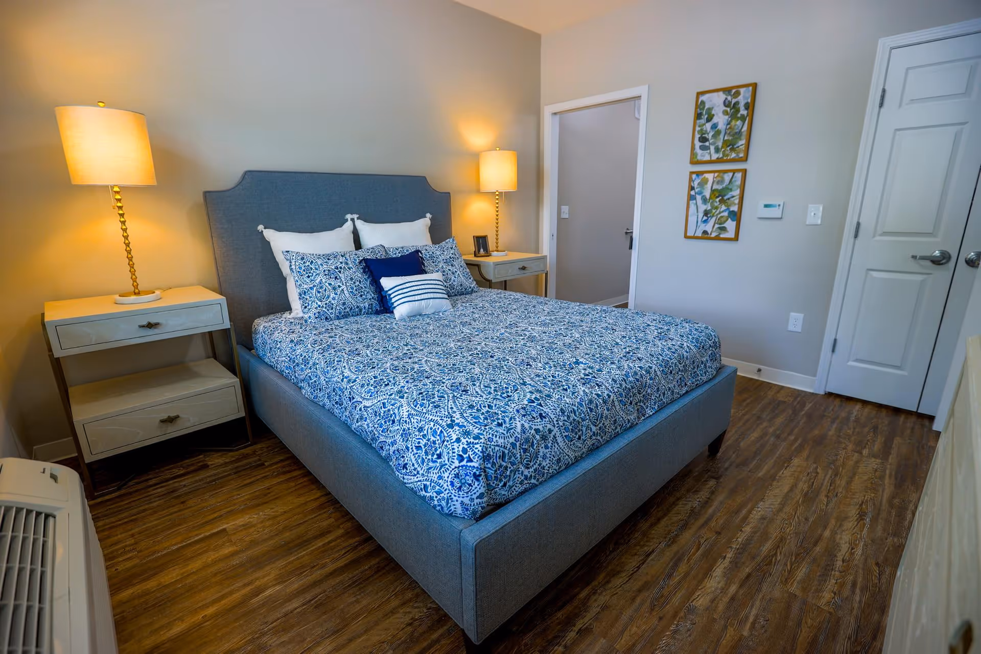 A neatly made bed with a blue patterned bedspread and multiple pillows in a bedroom. There are two nightstands on either side of the bed, each with a lit lamp. The room has wooden flooring, light-colored walls, two framed botanical prints on the wall, and a closed white door.