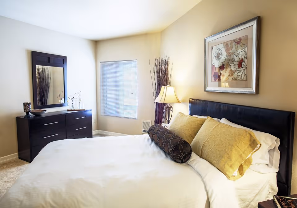 Bright furnished bedroom with a made bed, decorative pillows, a dark dresser and mirror, lamp, and framed artwork on the wall.