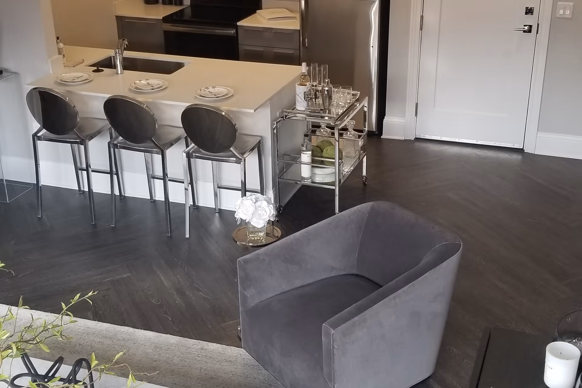 Modern interior space featuring a kitchen island with three metal bar stools, a small bar cart with glassware and bottles, a gray upholstered armchair, and a door in the background. The floor is dark wood with a herringbone pattern.