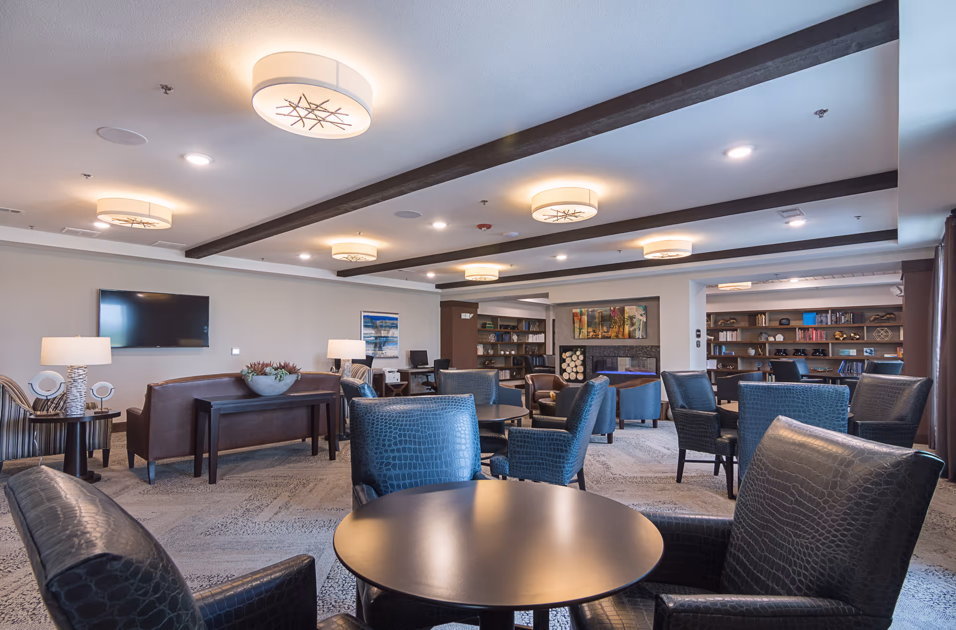 A spacious and well-lit common lounge area with multiple round tables surrounded by black leather chairs. The room features modern ceiling lights, a wall-mounted TV, a fireplace with stacked logs, bookshelves filled with books and decorative items, and a few table lamps. The decor is contemporary with neutral tones and wood accents.