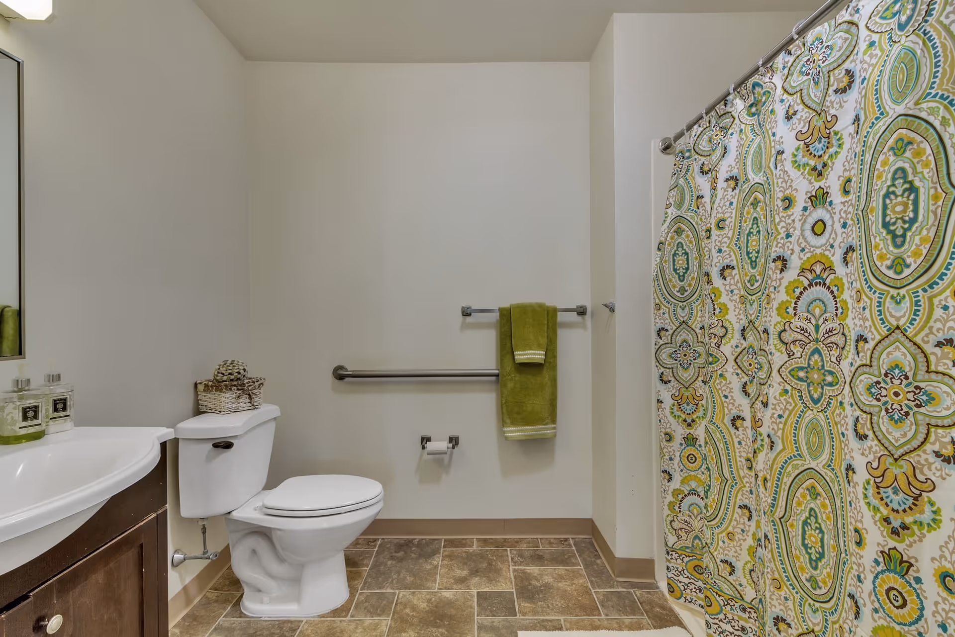 A clean bathroom with a white toilet, a sink with a dark wooden cabinet, a large mirror, and a shower with a colorful patterned curtain. There is a green towel hanging on a metal bar on the wall and a basket with toiletries on top of the toilet tank. The floor has brown and beige tiles.