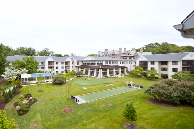 A large senior living facility building surrounding a spacious green courtyard with well-maintained lawns, trees, and shrubs. Several people are seen walking and sitting on benches in the courtyard area. The building has multiple floors with balconies and large windows.