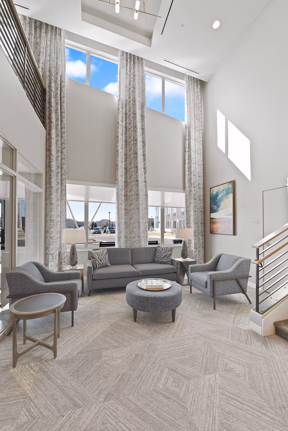 Bright two-story lobby seating area with tall windows, gray sofa and armchairs arranged around a round ottoman.
